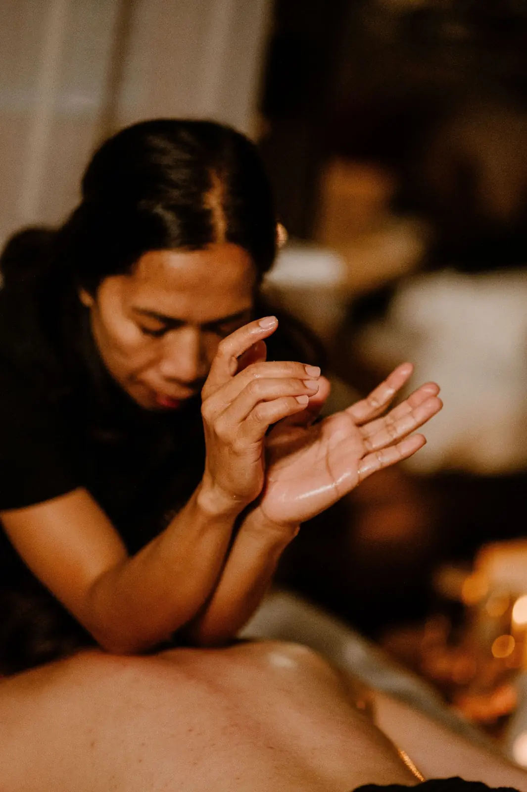 A Tera Thai therapist warms oil in her hands above a guest's back, with candles behind